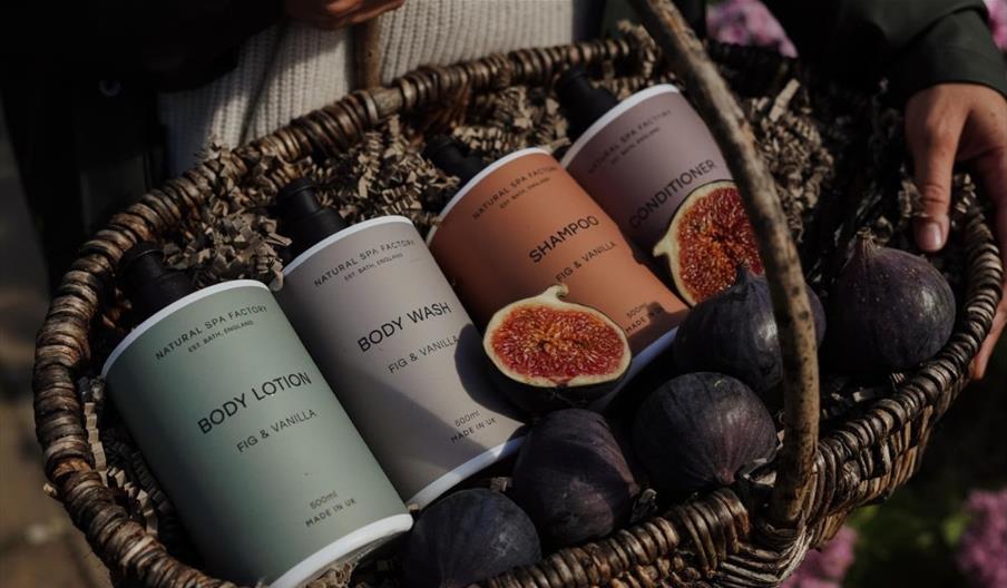 A woven basket containing figs and a selection of Natural Spa Factory branded toiletries.