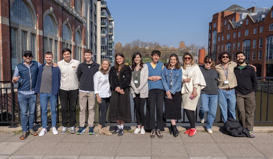 13 people in a row standing on a pavement with buildings and blue skies behind them.