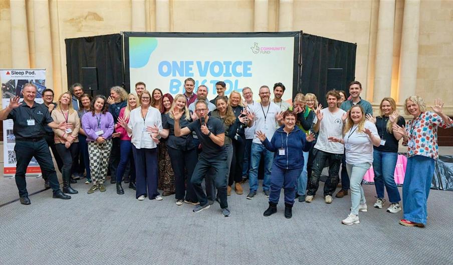 A large group of people with their hands raised in a jazz hands gesture, stood in front of a sign reading "One Voice Bristol".