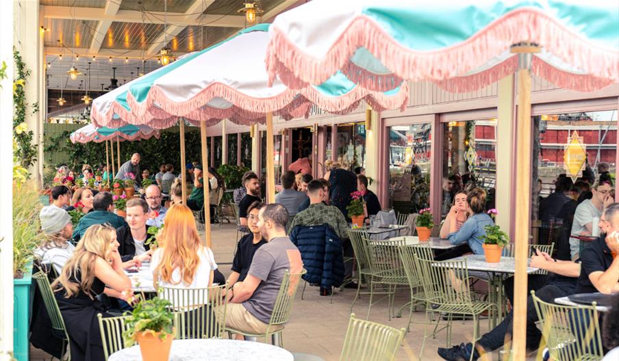 A large number of people seated in the outside dining area of Ritorno Lounge under pink and blue parasols.