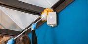 A lady dusting behind a speaker attached to a blue wall with a long duster