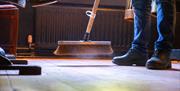 Someones feet and floor in shot with the bottom of a broom sweeping up dust