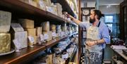 An employee wearing an apron decorated with a cow reaches towards a selection of cheeses on a shelf.