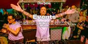 A DJ stood with his arms outstretched while standing in front of a mixing deck at a tiki-inspired event. White text describes Duchess' events services