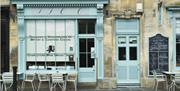 The exterior of Fine Cheese Co with a light blue door and window frames, hand-written blackboard menu, and outdoor seating.