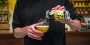 A man in front of a bar in a black t-shirt pouring a can of beer into a glass