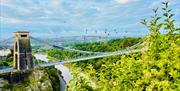 View of Clifton suspension bridge from above going over a gorge and river with lots of greenery surrounding it, blue skies with white clouds and a doz