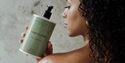 A woman having a shower, holding a bottle of Natural Spa Factory conditioner over her shoulder.