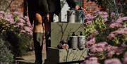 A person whose upper half is out of frame, standing in a garden of purple flowers beside a stack of boxes and Natural Spa Factory products.
