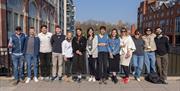 13 people in a row standing on a pavement with buildings and blue skies behind them.
