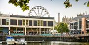 A view of the Ritorno Lounge building with the harbour in front. Two boats are docked on the harbour, and a ferris wheel is visible behind the buildin