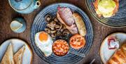 A plate of fried English breakfast with bacon, mushrooms, egg, sausage, beans and tomato, surrounded by side plates of other breakfast items.