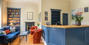 The reception at The Rodney Hotel with a blue desk and matching sofa, coffee table and bookcase on the left. Two orange armchairs face the sofa.