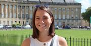 A photo of a young lady with dark bobbed hair and a smart white vest top, with the Bath Royal Crescent in the background on a sunny day.