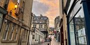 A crop of a cobbled Bath street with shops and old Cotswold stone buildings either side, and a pink sky in the background.